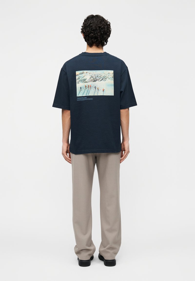 Mörkblå t-shirt i bomull med ett stort grafiskt tryck på ryggen som föreställer ett snötäckt landskap och figurer. Lös passform med korta ärmar.