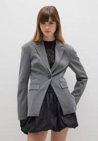 Grijze op maat gemaakte blazer, met enkele knoopsluiting, notch-lapels, voorkeuzepockets, gedragen over een zwarte kanten top en een zwarte uitlopende rok. Gladde textuur.
