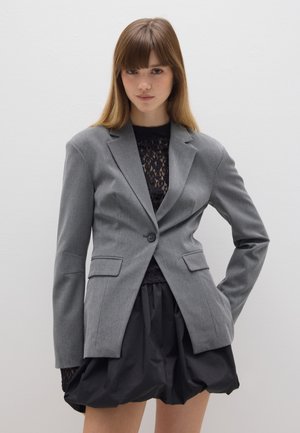 Grau maßgeschneiderter Blazer mit Knopfverschluss, Reverskragen, vorderen Taschen, getragen über einem schwarzen Spitzenoberteil und einem schwarzen Puffrock. Glatte Textur.