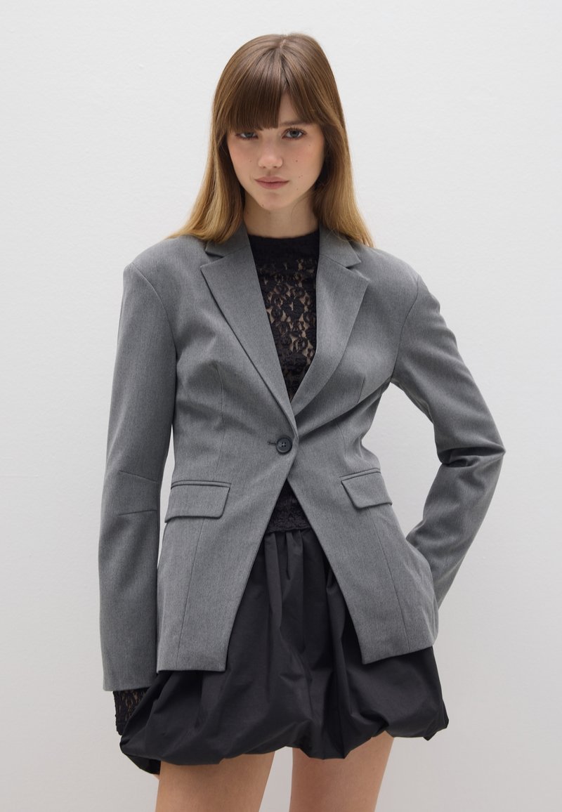 Grijze op maat gemaakte blazer, met enkele knoopsluiting, notch-lapels, voorkeuzepockets, gedragen over een zwarte kanten top en een zwarte uitlopende rok. Gladde textuur.