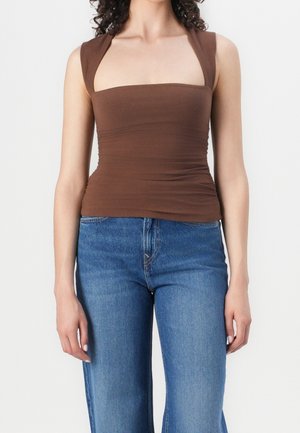 Vrouw die een nauwsluitend bruin mouwloos topje met brede bandjes en blauwe high-waisted jeans draagt, tegen een eenvoudige lichte achtergrond.