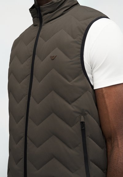 Άνδρας που φοράει ελιά πράσινο καπιτονέ γιλέκο με μαύρο φερμουάρ και λογότυπο πάνω από λευκό κοντομάνικο πουκάμισο, κροπαρισμένη άποψη του κορμού και του μπράτσου.