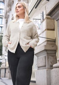 Ljusbeige kofta med en mjuk, texturerad yta, med framknappar och sidofickor, kombinerad med högmidjade svarta byxor.