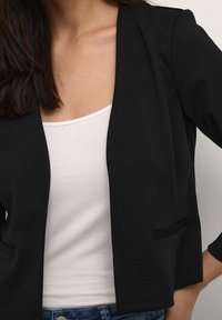 Svart kavaj med en mjuk textur, öppen framdesign och två framfickor, lager på lager över en vit linne. Avslappnade denimjeans synliga.