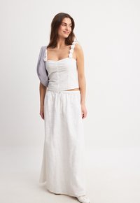 Biely crop top z očkovaného materiálu s nariasením, spárovaný s bielou maxi sukňou. Obsahuje volánové ramienka a fialový prekryv na jednom pleci.