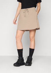 Falda corta beis con cinturilla elástica y cordón ajustable. Textura suave, hasta la rodilla, combinada con zapatos y calcetas negras.