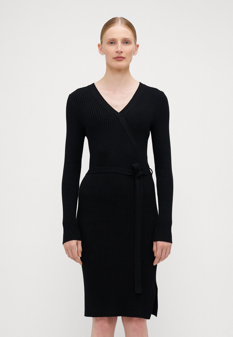 Sort strikket kjole med V-neck, lange ermer og belte i livet. Har ribbestrikket stoff og en splitt i siden i kanten.