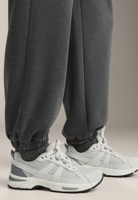 Világosszürke atlétikai cipők hálós és gumis részekkel, mintás talppal, sötétszürke felhajtott szárú nadrággal párosítva, laza stílust tükrözve.