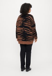 Cardigan tricoté brun et noir avec un motif en rayures de tigre, à manches longues et coupe décontractée, associé à un pantalon noir et des chaussures à plateforme.