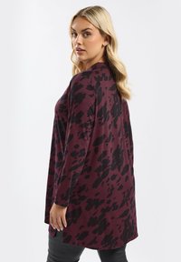 Una donna con i capelli biondi che indossa un cardigan a maniche lunghe color rosso porpora e nero con un motivo, mentre guarda oltre la sua spalla contro uno sfondo bianco.