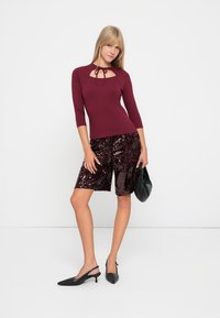 Bordowy dopasowany top z detalem w kształcie dziurki, w połączeniu z błyszczącymi czarnymi szortami z cekinami i czarnymi butami na obcasie typu slingback, uzupełniony małą czarną torebką.