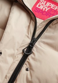 Beige gewatteerde jas met een zwarte ritssluiting, voorzien van een cirkelvormige trekker en een felroze merklabel met de opdruk "SUPERDRY SPORT."