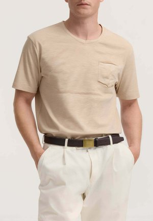 Uomo che indossa una maglietta beige a maniche corte con scollo a V e taschino sul petto, pantaloni bianchi e cintura scura con fibbia dorata, mani nelle tasche.