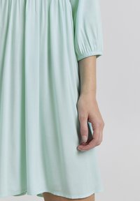Rochie verde mentă deschisă, cu decolteu rotund, talie strânsă, și mâneci 3/4 cu manșete elastice; țesătură netedă și fluidă.