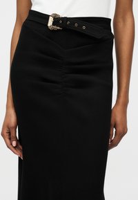 Mujer vistiendo una falda negra de talle alto con detalle fruncido en la parte delantera y un cinturón negro con una hebilla dorada ornamentada.