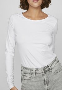 Långärmad vit t-shirt i slät tyg; har en rund halsringning och en figursydd design, parat med grå högmidjade jeans.