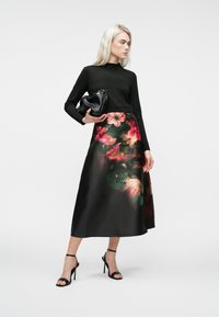 Žena nosi crnu bluzicu dugih rukava, cvjetnu midi suknju, crne sandale na visoku petu i drži crnu clutch torbicu, stoji protiv bijele pozadine.
