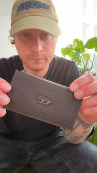 Szara skórzana portmonetka na zamek z fakturowanym wykończeniem, owalnym logo jako ozdobą, trzymana przez osobę. W tle zielona roślina i miękkie oświetlenie.