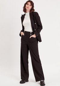 Schwarze, taillierte Blazerjacke mit silbernen Knöpfen, weißem geripptem Oberteil und weit geschnittenen schwarzen Hosen, die seitliche Taschen und klobige schwarze Schuhe aufweisen.
