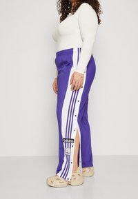 Fialové sportovní kalhoty s bílými bočními pruhy a patentkami. Vyrobené z tkaniny s hladkou texturou, s nášivkou loga Adidas na boku.