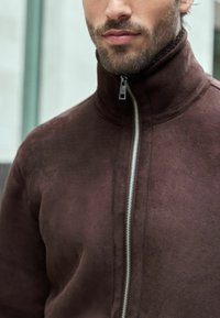 Jachetă din suede maro cu guler înalt și fermoar argintiu. Textura pare moale, având detalii de design minimaliste.