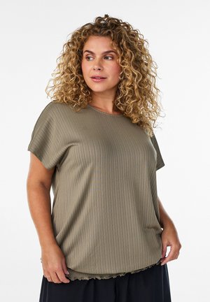 Žena s kučeravými vlasmi, oblečená v voľnej, rebrovanej, olivovo zelenej krátkym rukávom a čiernej sukni, pozrite si mierne do strany na bielom pozadí.