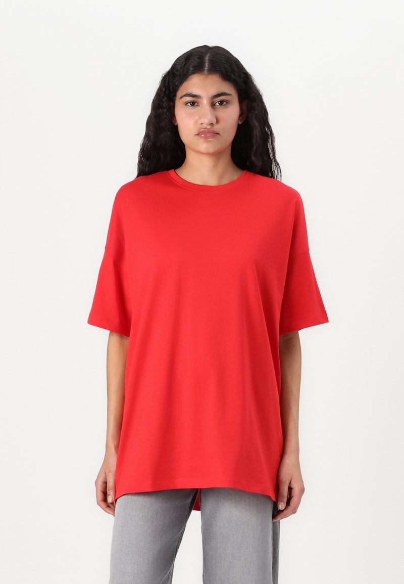 Red cotton t-shirt with short sleeves, crew neck, and a relaxed fit. Smooth texture and straight hem, worn with light gray trousers.