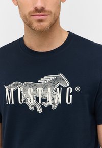 Tumši zilas krāsas T-krekls, kas izgatavots no kokvilnas, ar baltu grafiku, kurā attēlots skraidošs zirdziņš un vārds "MUSTANG" treknā, stilizētā fontā.