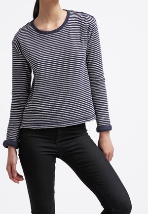 Persoon draagt een navy-wit gestreept longsleeve met opgerolde manchetten en zwarte aansluitende broek tegen een effen witte achtergrond.