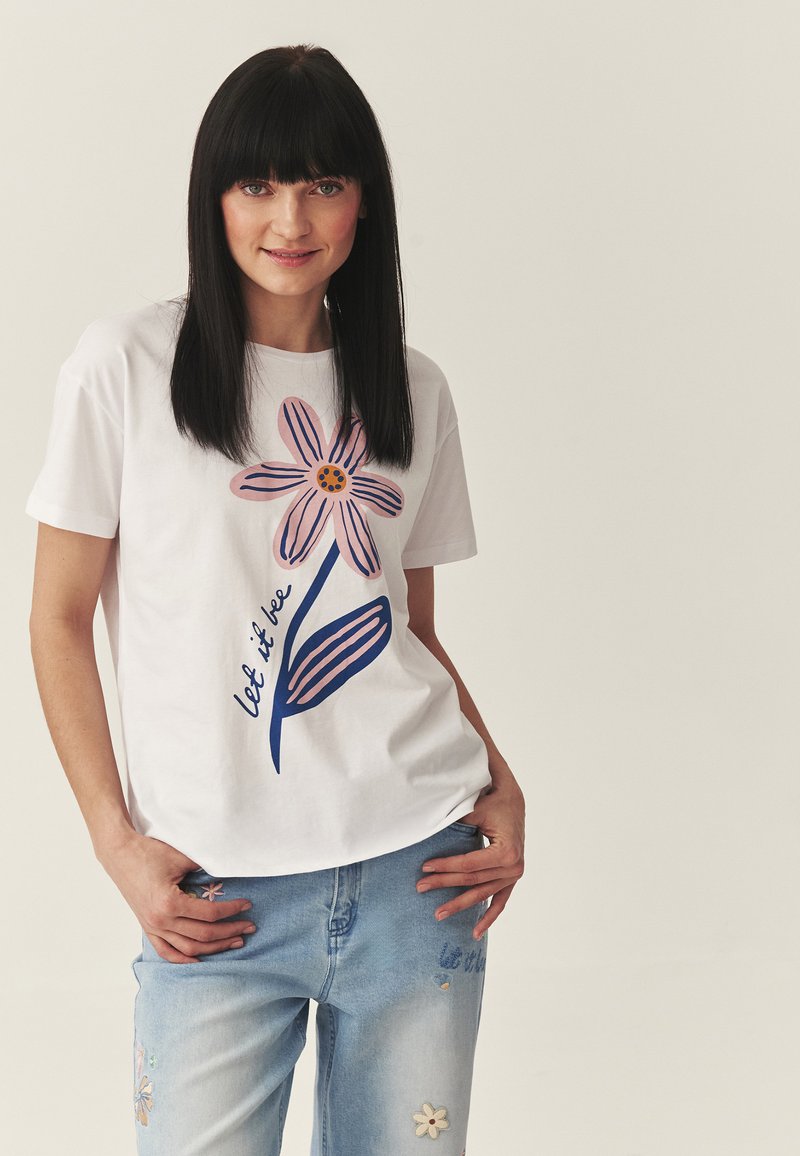 Femme aux cheveux noirs lisses portant un t-shirt blanc avec une grande fleur rose et le texte « let it be », assorti à un jean bleu avec une broderie florale.
