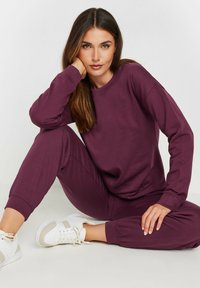 Sudadera burdeos y joggers a juego en una tela suave, con mangas largas, puños de canalé y un ajuste relajado. Combinados con zapatillas blancas.