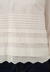 Blouse blanche avec plis horizontaux au-dessus d'un ourlet festonné avec découpes de fleurs ajourées, associée à un jean denim bleu foncé.
