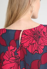 Floral blouse featuring a navy blue fabric with large red flowers, a back keyhole cutout, and a decorative button accent.