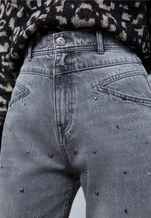 Harsoina, korkeavyötäröisinä harmaina farkuilla, joissa on hajallaan metallinappeja, paritettu musta-beige kuvioituneen neuleen kanssa, joka on työnnetty sisään.