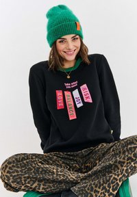 Černá mikina s barevným grafickým nápisem "Vezmi si, co potřebuješ" a slovy: LÁSKA, ROVNOVÁHA, RADOST, RIZIKO. Kalhoty s leopardím vzorem a zelená čepice.