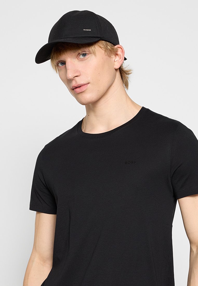 Junger Mann, der eine schwarze Mütze mit dem Text "SANDRO" und ein schwarzes BOSS-T-Shirt trägt, schaut leicht zur Seite vor einem schlichten Hintergrund.