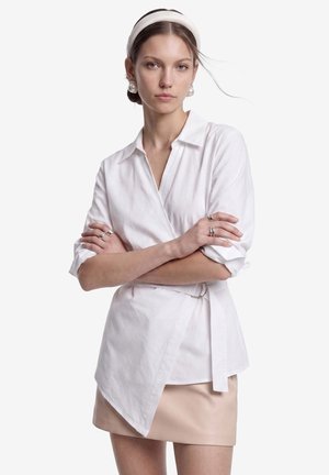Joven mujer con cinta blanca en la cabeza y pendientes de perlas que lleva una blusa cruzada blanca y una falda beige, con los brazos cruzados, expresión neutra, de pie contra un fondo blanco.