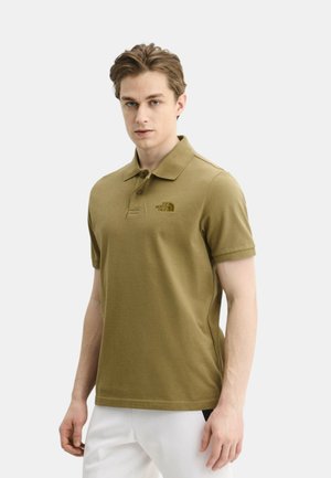 Młody mężczyzna w oliwkowej koszulce polo z logo The North Face i białych spodniach, stojący na jednolitym jasnym tle.