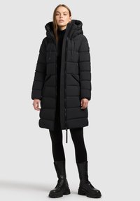 Schwarzer Steppmantel mit Kapuze, mit gesteppter Optik, Seitentaschen und durchgehendem Frontreißverschluss. Kombiniert mit schwarzen Leggings und klobigen Stiefeln.