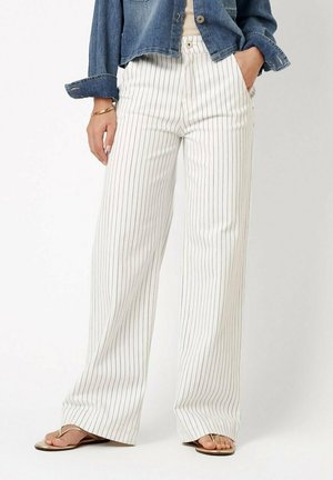 Woman wearing white wide-leg pants with thin black vertical stripes, denim jacket, gold bracelet, and gold sandals, standing against a white background.