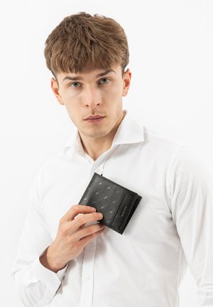 Jonge man in wit overhemd houdt een zwarte leren portemonnee met reliëfpatronen dicht bij zijn borst vast, kijkt recht vooruit.