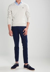 Pull léger gris clair en V, chemise rayée bleu clair en dessous, pantalon slim-fit bleu marine, et chaussures en daim bleu foncé avec semelles blanches.