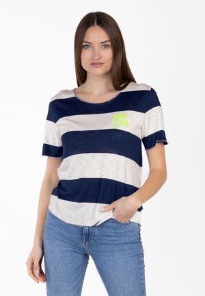 Kobieta w granatowo-białej koszulce w paski z neonowym zielonym nadrukiem palmy, w niebieskich dżinsach, stoi z jedną ręką w kieszeni.