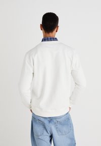 Sudadera blanca de manga larga, con cuello redondo y puños de canalé. Lleva una camisa de cuadro azul debajo, combinada con unos vaqueros de un azul claro.