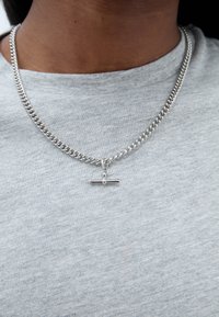 Strieborný náhrdelník s reťazou so širokým článkovým dizajnom a horizontálnym barovým príveskom, nosený cez šedý textilný top. Hladký povrch a kovový lesk.