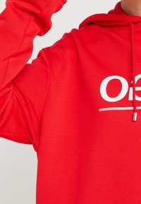 Punainen huppari, jossa on sileä pinta, suuri valkoinen logo ja metallipäällysteinen kiristysnaru. Löysä ja rento malli.