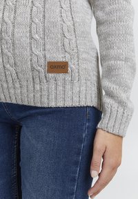 Lys grå strikkegenser med kabel- og ribbestrikkede mønstre. Har et brunt lærmerke med «oxmo»-tekst. Båret over blå jeans.