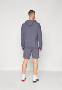 Grå bomulds hættetrøje med snoretræk i huen, parret med matchende shorts. Båret med hvide sneakers med sorte og røde detaljer.