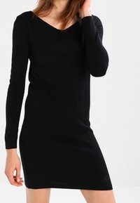 Schwarzes gestricktes Kleid mit V-Ausschnitt, langen Ärmeln und figurbetonter Silhouette. Bietet eine glatte Textur und dezente Markierung nahe dem Saum.