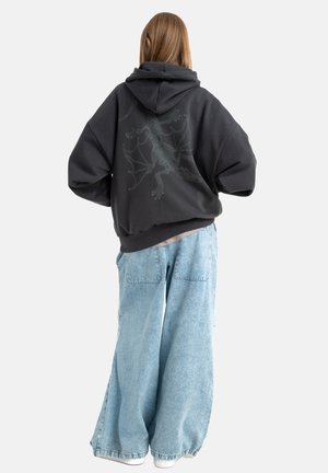 Czarna, oversize bluza z kapturem z grafiką szkieletonu smoka na plecach, zestawiona z jasnoniebieskimi dżinsami o szerokich nogawkach. Bawełnowy materiał, luźny krój.